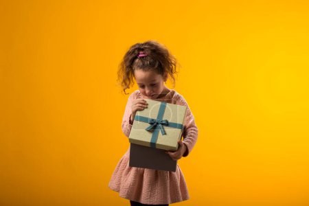 Téléchargez les photos : Portrait d'une jeune fille souriante tenant une boîte-cadeau et regardant à l'intérieur de la boîte sur fond jaune. Concept d'anniversaire et de célébration - en image libre de droit