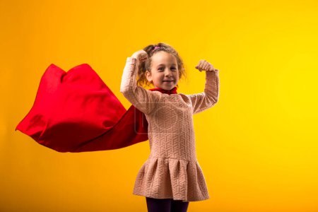 Portait of kid girl superhero in a red cloak showing strenght gesture on yellow background. Concept of victory and success.