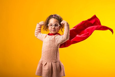 Foto de Portait de niña superhéroe en una capa roja que muestra gesto de fuerza sobre fondo amarillo. Concepto de victoria y éxito. - Imagen libre de derechos