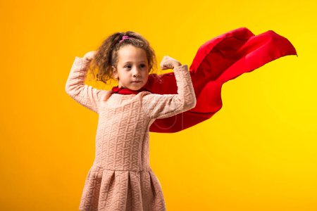 Portait of kid girl superhero in a red cloak showing strenght gesture on yellow background. Concept of victory and success.