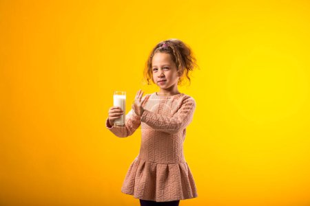 Téléchargez les photos : Portrait d'une fillette allergique aux produits laitiers tenant un verre de lait sur fond jaune. Concept d'intolérance au lactose - en image libre de droit