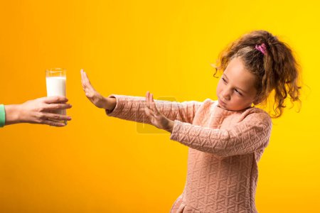 Téléchargez les photos : Portrait de gamine allergique aux produits laitiers abandonnant le verre de lait sur fond jaune. Concept d'intolérance au lactose - en image libre de droit