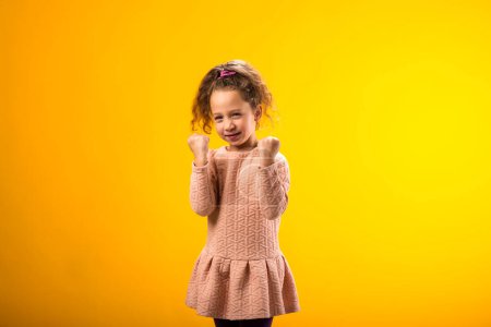 Portrait of smiling kid girl showing success over yellow background. Luck concept