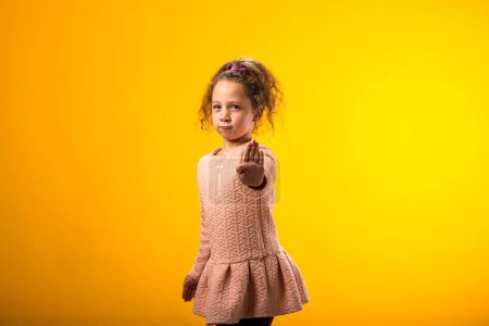 Portrait of child girl showing stop gesture on yellow background. Bulling and negative emotions concept