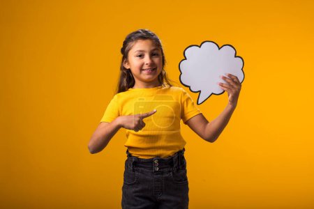 Foto de Retrato de una niña sonriente sosteniendo una tarjeta de burbuja de nube y señalándola con el dedo sobre un fondo amarillo. Concepto de ensueño - Imagen libre de derechos