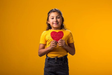 Téléchargez les photos : Portrait d'une enfant fille tenant papier heartover fond jaune. Amour, soins et concept de Saint Valentin - en image libre de droit
