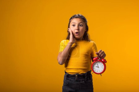 Photo for Portrait of surprised kid girl holding an alarm clock in hand. The concept of education, school, deadlines, time to study - Royalty Free Image