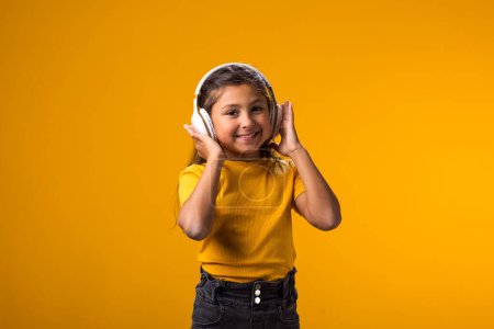 Téléchargez les photos : Portrait d'une jeune fille souriante portant une robe rose jouissant de la musique dans un casque sur fond jaune. Concept de style de vie et de loisirs - en image libre de droit