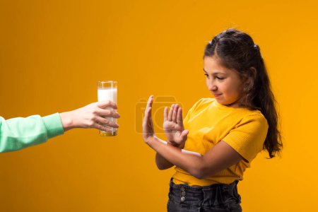 Téléchargez les photos : Portrait d'une fillette allergique aux produits laitiers montrant un geste d'arrêt devant un verre de lait sur fond jaune. Concept d'intolérance au lactose - en image libre de droit