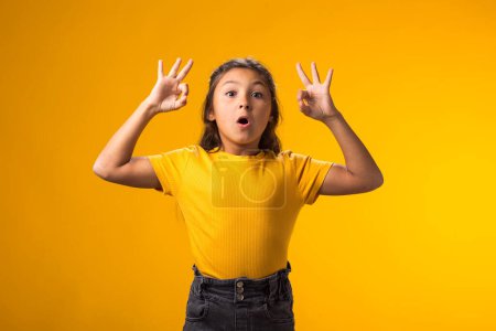 Téléchargez les photos : Portrait d'une enfant surprise regardant à travers un geste correct sur fond jaune. Concept des émotions positives - en image libre de droit