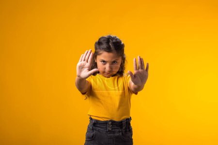 Portrait of child girl showing stop gesture on yellow background. Bulling and negative emotions concept