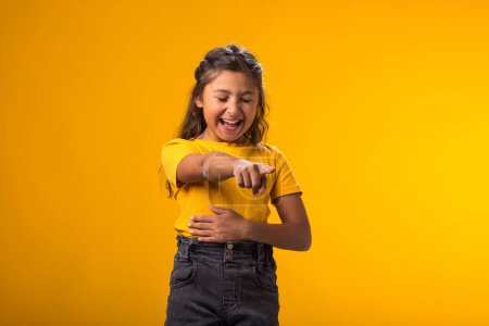 Retrato de niña burlándose y burlándose de alguien mostrando el dedo en la cámara y sosteniendo el estómago sobre el fondo amarillo. Concepto Bulling