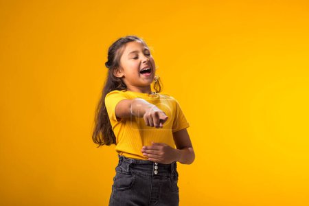 Portrait of kid girl mocking and teasing at someone showing finger at camera and holding stomach over yellow background. Bulling concept