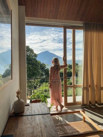 A middle-aged woman in red dress stands near the balcony door and looks at the jungle and mountains on a sunny summer day