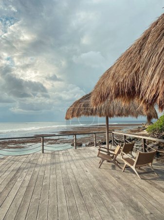 Wooden deck with straw beach umbrellas, chairs and sunset view in Uluwatu cliff, Bukit Peninsula, Bali.