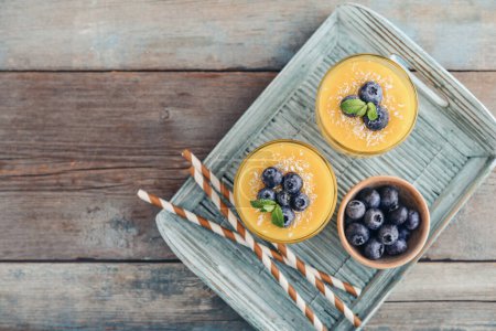 Erfrischender und gesunder Mango-Smoothie in Gläsern mit Kokosflocken und frischen Blaubeeren auf Holzgrund, Draufsicht