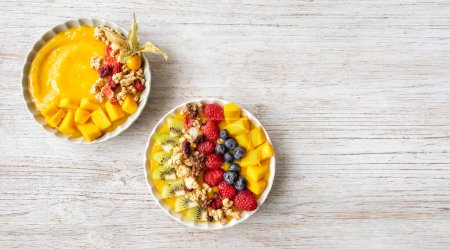 Photo for Ywo Healthy mango smoothie bowls with blueberries, raspberries, kiwi and granola. Above view scene on a light grey  wooden background. - Royalty Free Image