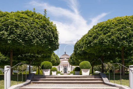 A lush garden pathway leading to a pavilion in Melk Abbey, Austria, surrounded by trimmed bushes and symmetrical trees under a sunny sky.