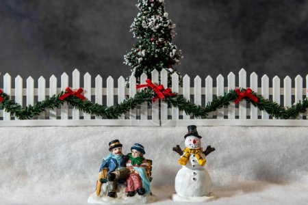Foto de Escena horizontal de Navidad con figuras de marido y mujer sentados en el banco del parque en la nieve junto al muñeco de nieve - Imagen libre de derechos