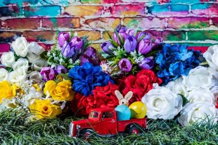 Foto de Red toy truck in green grass with colored Easter eggs and bunny surrounded by spring flowers and colorful brick wall background - Imagen libre de derechos
