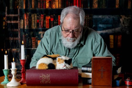 Foto de Senior caucásico macho lectura biblia en retro buscando biblioteca por vela con calico gato - Imagen libre de derechos
