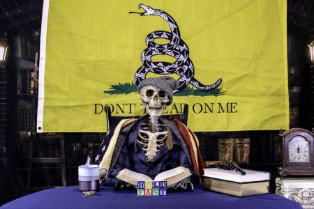 Foto de Esqueleto con sombrero y bufanda libro de lectura en la biblioteca con banderas estadounidenses y Gadsden con pistola y bloques ortografía aferrarse rápidamente - Imagen libre de derechos