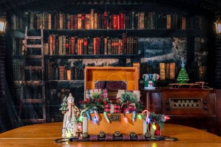 Foto de Maleta vintage en mesa de roble con radio antigua y fondo de biblioteca vieja llena de regalos y decoraciones de Navidad - Imagen libre de derechos