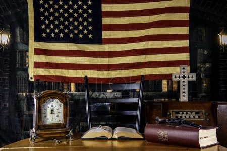 Pistole und Bibel auf Holztisch in alter Bibliothek mit Vintage-Radio und amerikanischer Flagge