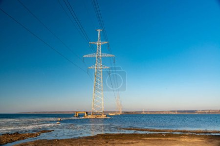 Foto de High-voltage power line against the background of the blue sky above the water of the lake - Imagen libre de derechos