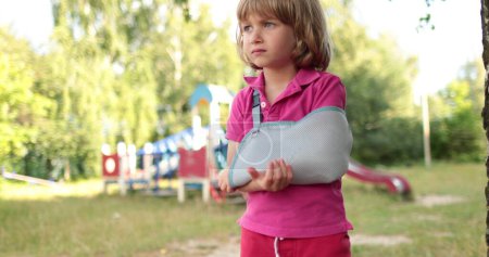Téléchargez les photos : Un petit, beau garçon avec un bras cassé enveloppé dans un bandage. Un enfant blessé sur le terrain de jeu. - en image libre de droit
