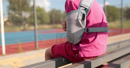Foto de Niño con la extremidad rota al aire libre. un niño con un brazo roto se sienta cerca del campo de deportes. - Imagen libre de derechos