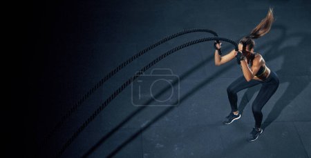 Efektywny trening z liną. Sportswoman trenuje na siłowni funkcjonalnej, wykonując ćwiczenia crossfit z liną bojową.
