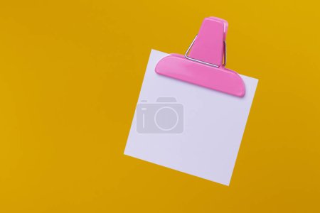 White paper note with pink paper clip holder isolated on yellow background. Blank note on yellow.