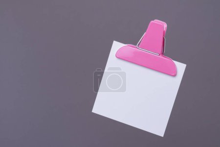 White paper note with pink paper clip holder isolated on gray background. Blank note on gray.