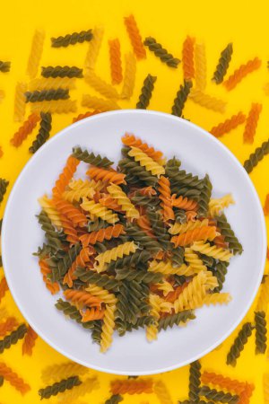 Three-colored uncooked pasta in the white bowl on yellow background with scattered pasta on it. Top view.