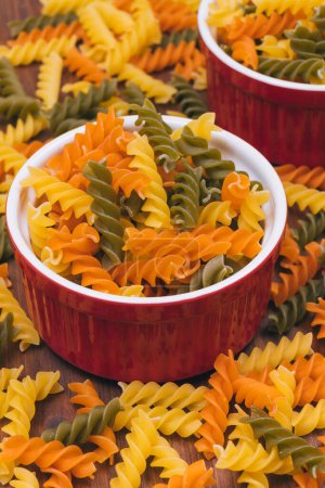 Three-colored uncooked pasta in the ceramic bowls on the wooden background with scattered pasta on it.