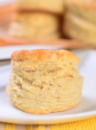 Photo for Freshly baked flaky biscuit served on a white plate with more biscuits in the background - Royalty Free Image