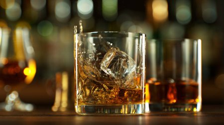 Foto de Ice cube fall into a glass with a golden alcoholic drink. Bar on background. - Imagen libre de derechos
