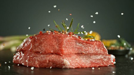 Foto de Primer plano de la caída de sal y pimienta en filetes de carne cruda en la mesa, congelar el movimiento. - Imagen libre de derechos