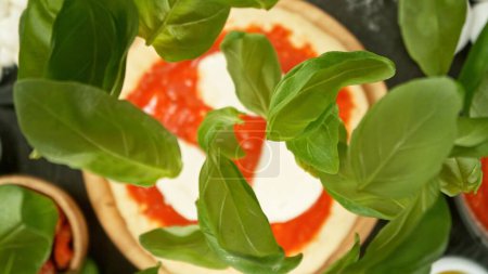 Foto de Freeze Motion of Falling Basil Leaves on Pizza Dough (en inglés). Vista superior de la mesa, concepto de preparación de alimentos, ángulo de visión único. - Imagen libre de derechos