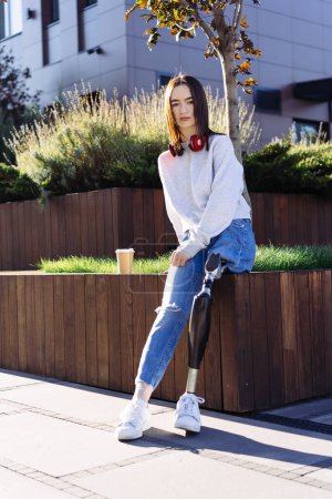 Photo for Female with prosthetic leg sitting in city. Woman with prosthetic leg wearing red headphones and drinking coffee. Woman with leg prosthesis equipment. - Royalty Free Image