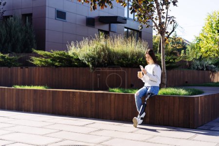 Photo for Female with prosthetic leg sitting in city. Disabled woman with prosthetic leg in red headphones, drinking coffee and using smartphone. Woman with leg prosthesis equipment. - Royalty Free Image