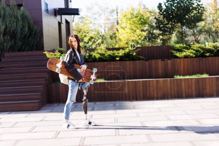 Photo for Portrait of young woman with prosthetic leg with longboard in city. Disabled woman student having a walk. Woman with leg prosthesis equipment. - Royalty Free Image