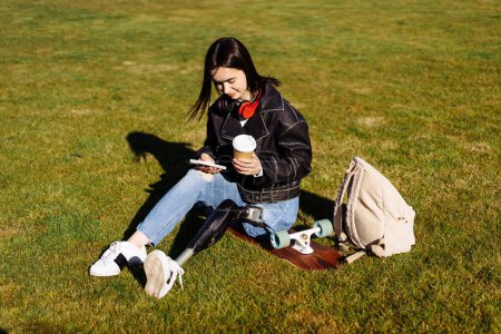Photo for Young woman student with prosthetic leg sitting on green grass in university campus. Disabled woman with bionic leg. Woman with leg prosthesis equipment using phone. - Royalty Free Image
