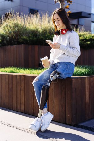 Photo for Female with prosthetic leg sitting in city. Woman with prosthetic leg wearing red headphones and drinking coffee. Woman with leg prosthesis equipment. - Royalty Free Image