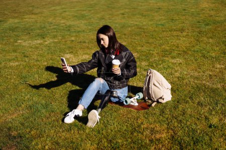 Photo for Young woman student with prosthetic leg sitting on green grass in university campus and making selfie with smartphone. Disabled woman with bionic leg. Woman with leg prosthesis equipment using phone. - Royalty Free Image