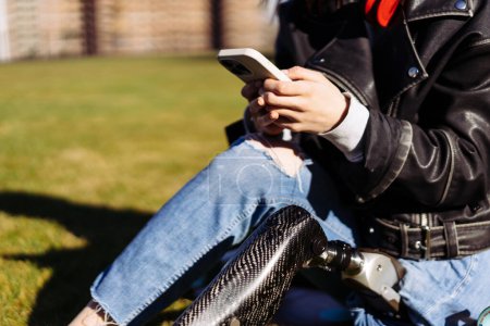 Photo for Close up of woman student with prosthetic leg sitting on green grass in university campus and using smartphone. Disabled people with bionic leg in park. Woman with prosthesis equipment using phone. - Royalty Free Image
