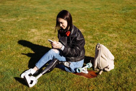 Photo for Young woman student with prosthetic leg sitting on green grass in university campus and using smartphone. Disabled woman with bionic leg in public park. Woman with prosthesis equipment using phone. - Royalty Free Image