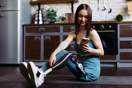 Photo for Lady with prosthetic limb sits on mat to relax after training. Woman with disability chats with close friends on social media after adaptive yoga session - Royalty Free Image