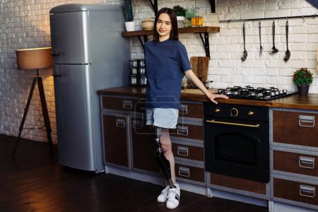 Photo for Brunette woman with artificial limb stands in kitchen with crossed arms. Female with disability exudes confidence posing for camera in cozy kitchen - Royalty Free Image